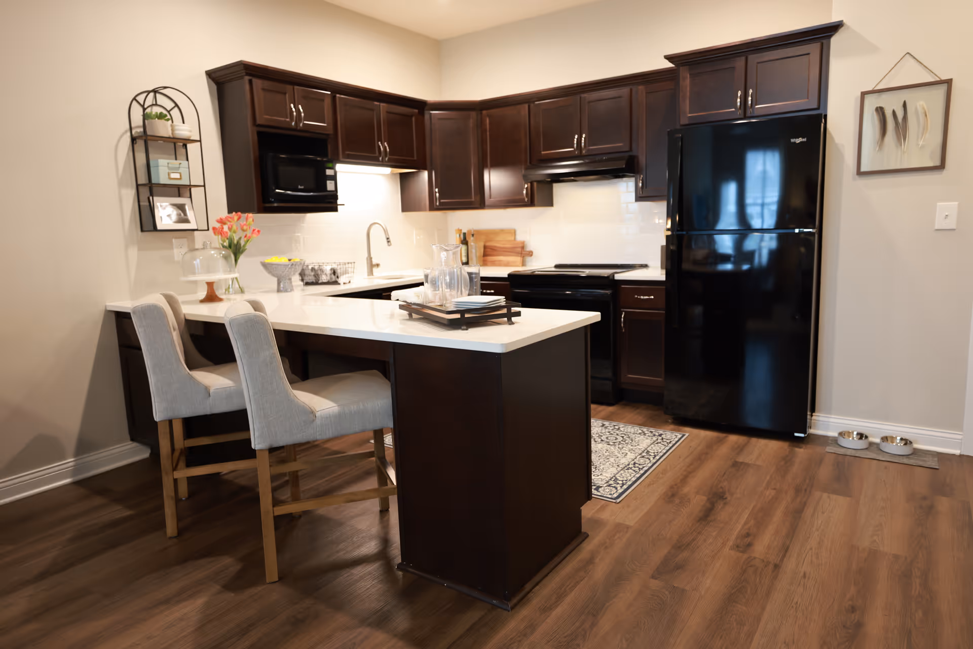 Modern kitchen with dark wood cabinets, a black refrigerator, black stove, and microwave. There is a white countertop island with two beige upholstered chairs. The floor is wood, and there are decorative items including flowers, a framed picture, and a small shelf on the wall.