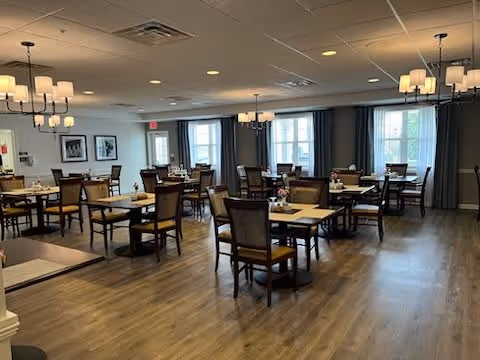 A spacious dining room with multiple wooden tables and chairs arranged neatly. The room has large windows with curtains allowing natural light to enter, and several modern chandeliers hanging from the ceiling. The floor is wooden, and the walls are decorated with framed pictures.