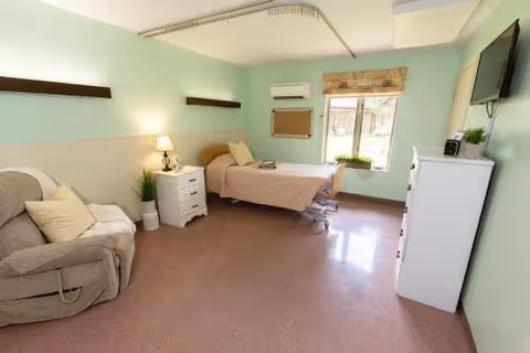 A bright and clean bedroom in a healthcare facility with a single hospital-style bed, a beige recliner chair with a pillow and blanket, a white nightstand with a lamp and plant, a window with blinds, a wall-mounted TV, and a white dresser with a small plant on top. The walls are painted light green and the floor is a smooth, light brown surface.