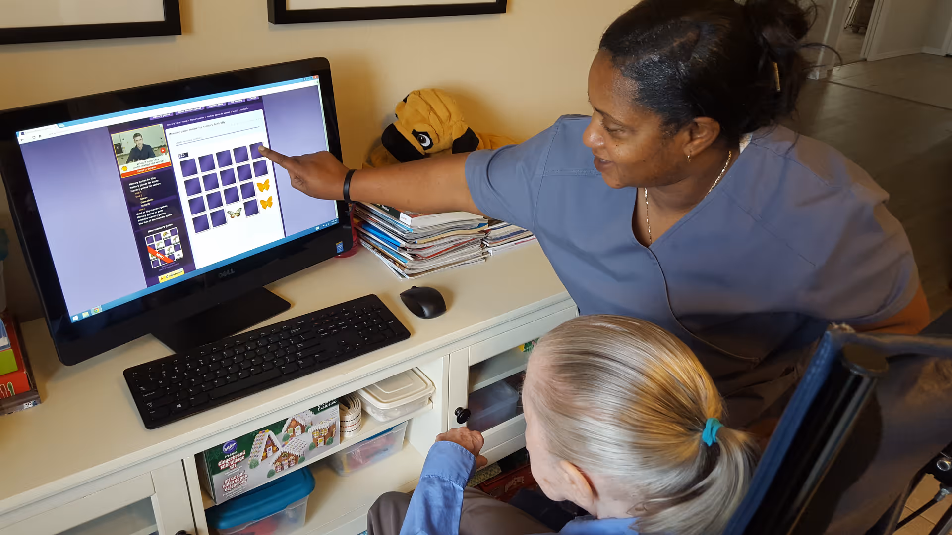 A caregiver in blue scrubs is assisting an elderly woman with gray hair tied in a ponytail, who is seated in a wheelchair, as they look at a computer screen displaying a memory or matching game with butterfly images.