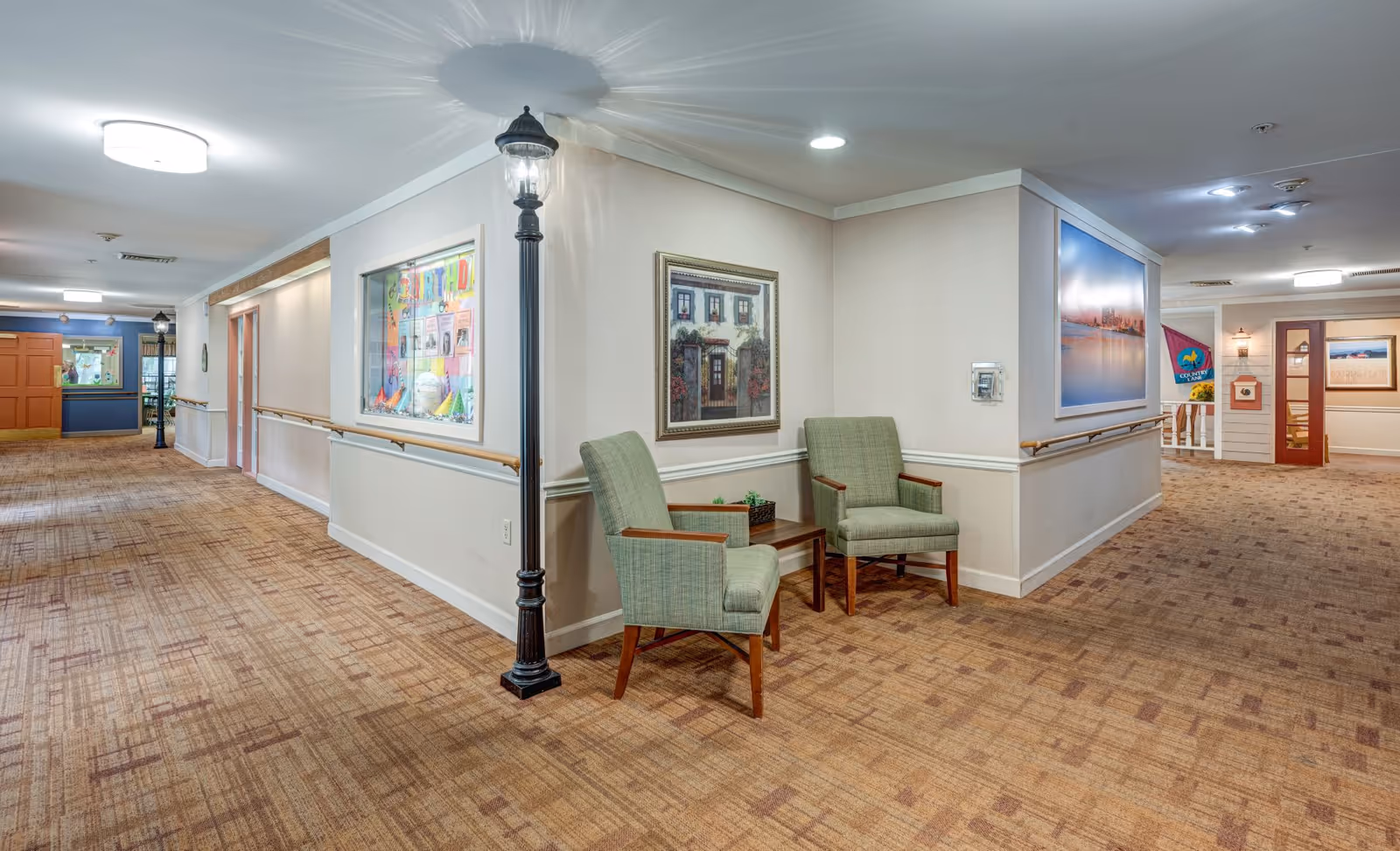 A well-lit hallway in a senior living facility with beige walls and patterned carpet. Two green upholstered chairs with wooden arms are placed against a corner wall with a framed painting above them. A decorative black street lamp stands next to the chairs. The hallway has handrails along the walls and various framed pictures and bulletin boards. Doors and additional seating areas are visible further down the hall.