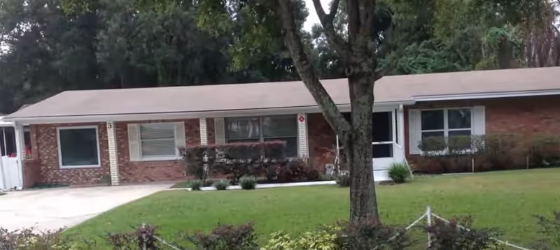 Front of a single-story brick senior living facility with windows, lawn, shrubs, and a tree.