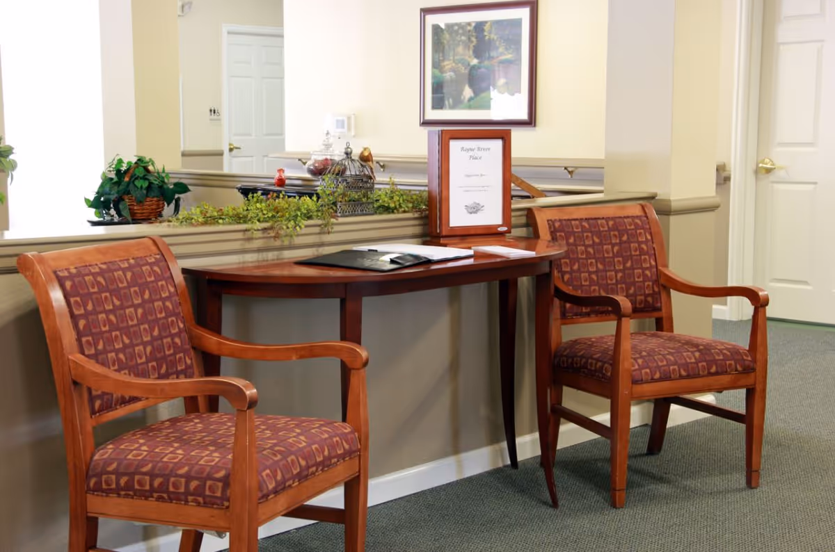 A small seating area with two wooden chairs featuring patterned upholstery and a wooden half-moon table between them. On the table are some documents and a framed sign that reads 'Rogue River Place'. The background shows a beige wall with a framed picture and two white doors.