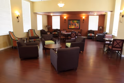 Communal lounge with grouped armchairs, small tables, and a dining table on a polished wood floor under warm lighting.