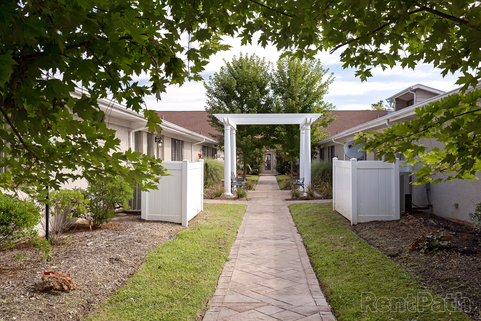 A paved walkway lined with grass and shrubs leads through a white pergola between two single-story buildings with beige brick walls and brown roofs. Trees and benches are visible along the path, and the scene is framed by green leaves from overhanging branches.