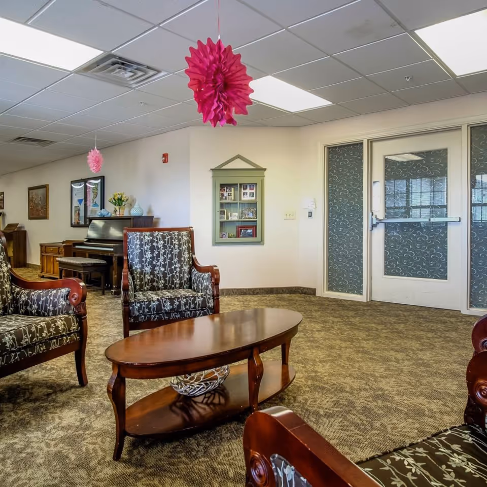 A cozy living room area in an assisted living facility featuring patterned armchairs around a wooden coffee table, a piano against the wall with decorative items on top, framed pictures on the walls, and two pink hanging paper decorations from the ceiling. There is a door with frosted glass panels on the right side.