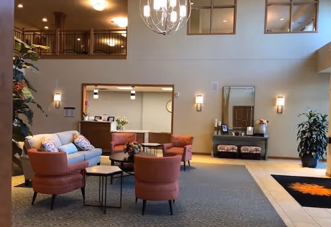 A spacious and well-lit senior living facility common area with a high ceiling and a chandelier. The room features a light blue sofa with patterned pillows, four coral-colored armchairs arranged around small tables, a reception desk in the background, a large mirror above a console table with two cushioned stools, and several potted plants. The upper level has a railing overlooking the area.