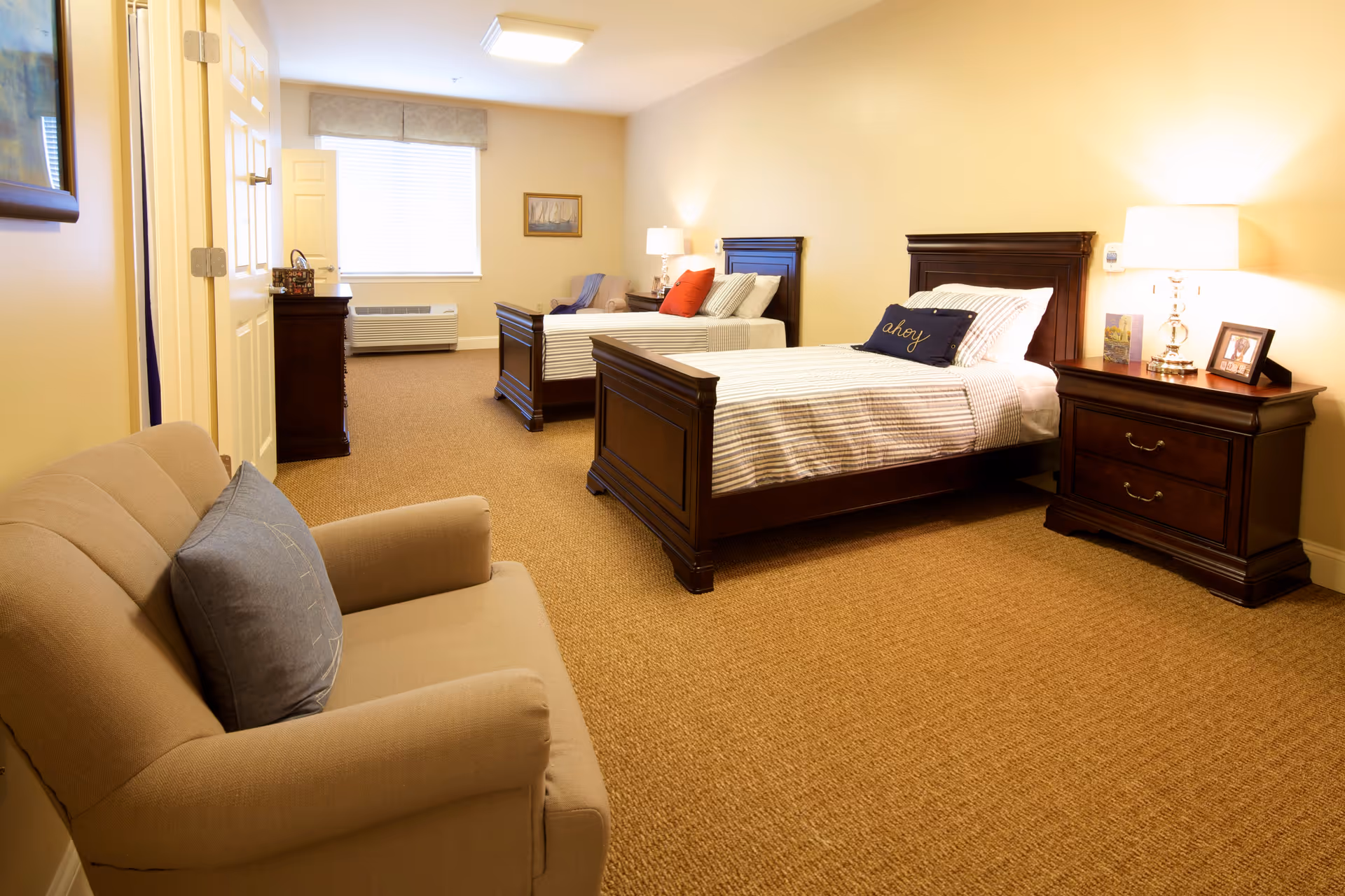 A warmly lit bedroom with two single beds featuring dark wooden frames and striped bedding. Each bed has a decorative pillow, one with the word 'ahoy'. There is a wooden nightstand with a lamp, framed photos, and a card between the beds. A beige armchair with a blue pillow is positioned in the foreground. A window with blinds and a valance is at the far end of the room, next to a dark wooden dresser. The walls are painted a soft yellow, and the floor is covered with a brown carpet.