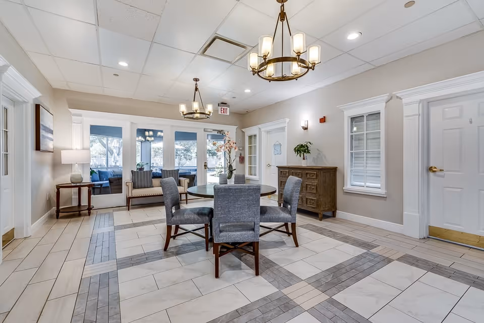A bright and spacious common area in a senior living facility featuring a round table with four upholstered chairs in the center. The room has tiled flooring with a decorative pattern, beige walls, and white trim. Two chandeliers hang from the ceiling, and there is a wooden sideboard with a plant on top against one wall. Large glass doors lead to an outdoor seating area with blue cushions. Additional seating includes a bench with cushions near the glass doors and a small side table with a lamp.