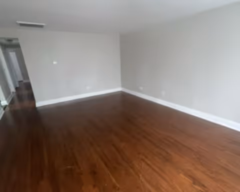 Empty room with wooden flooring, white walls, and a hallway leading to other rooms.
