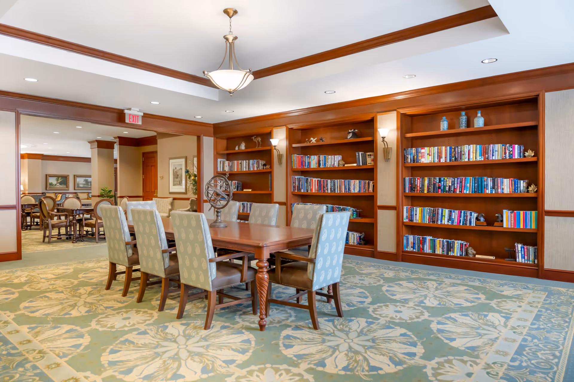 A well-lit interior room with a large wooden table surrounded by eight upholstered chairs. Behind the table are built-in wooden bookshelves filled with books and decorative items. The room has a patterned carpet and a chandelier hanging from the ceiling. In the background, there are more tables and chairs in an adjoining room.