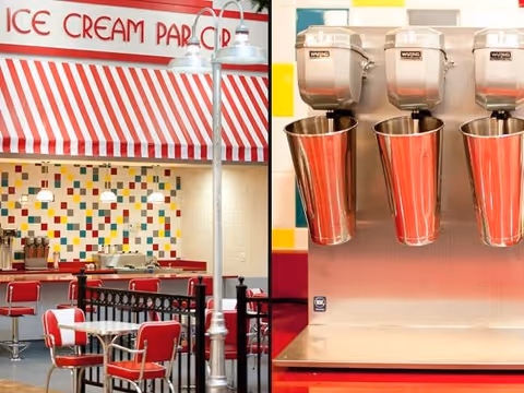 Interior view of an ice cream parlor with red and white striped awning, red chairs and tables, colorful tiled wall, and a close-up of three stainless steel milkshake mixers with metal cups attached.