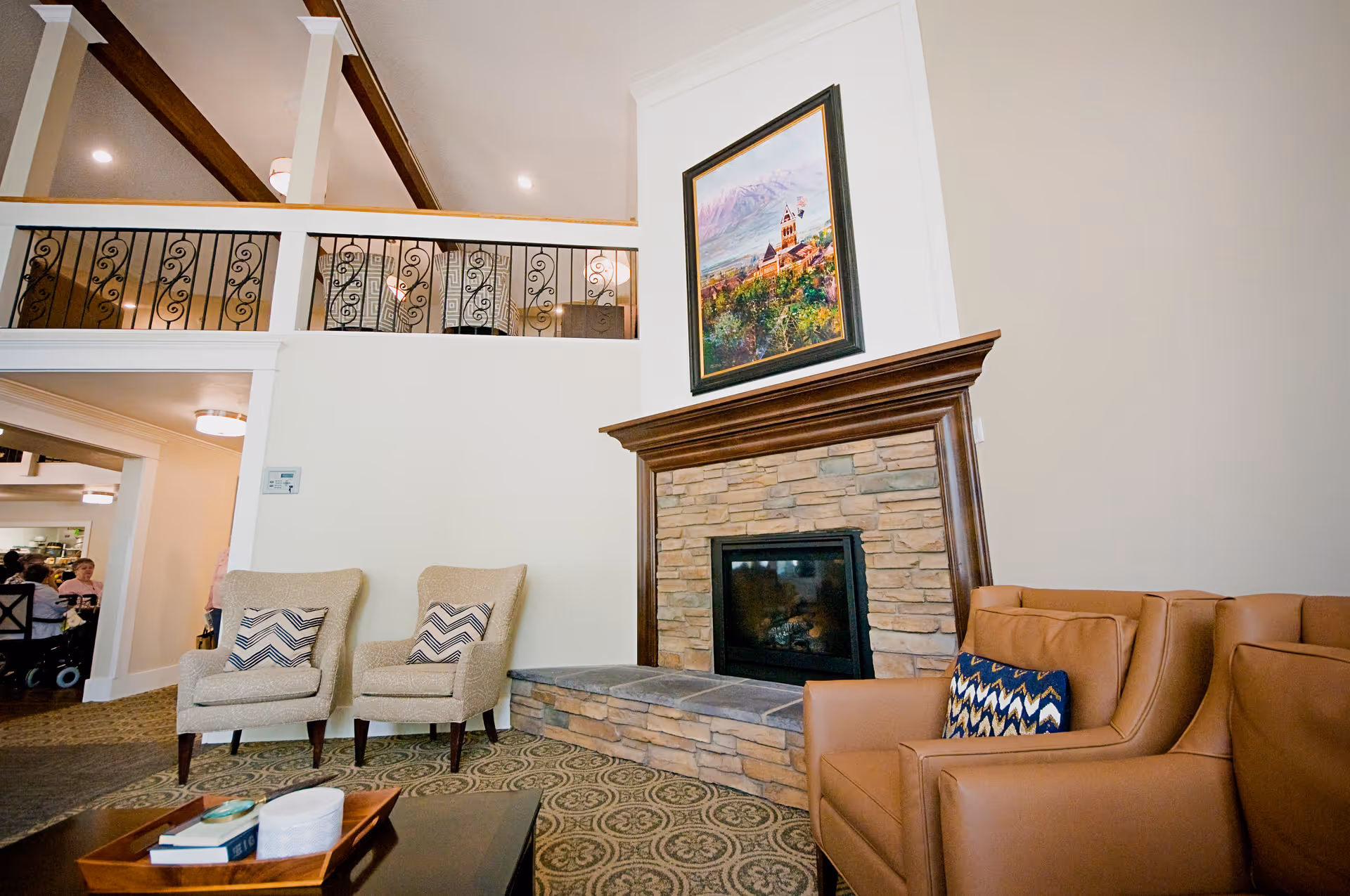 A cozy living room area in Williamsburg Retirement Community featuring a stone fireplace with a wooden mantle, a framed painting above it, two beige armchairs with patterned cushions, and a brown leather sofa with a blue patterned cushion. The room has a high ceiling with exposed beams and a decorative railing on the upper level.