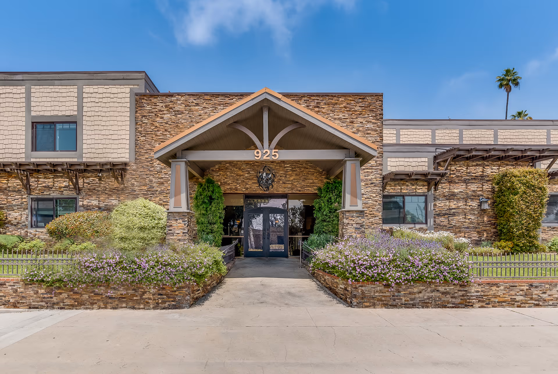 Front exterior view of Astoria Park Senior Living building with stone facade, a covered entrance with the number 925 above it, surrounded by landscaped bushes and flowers under a blue sky.