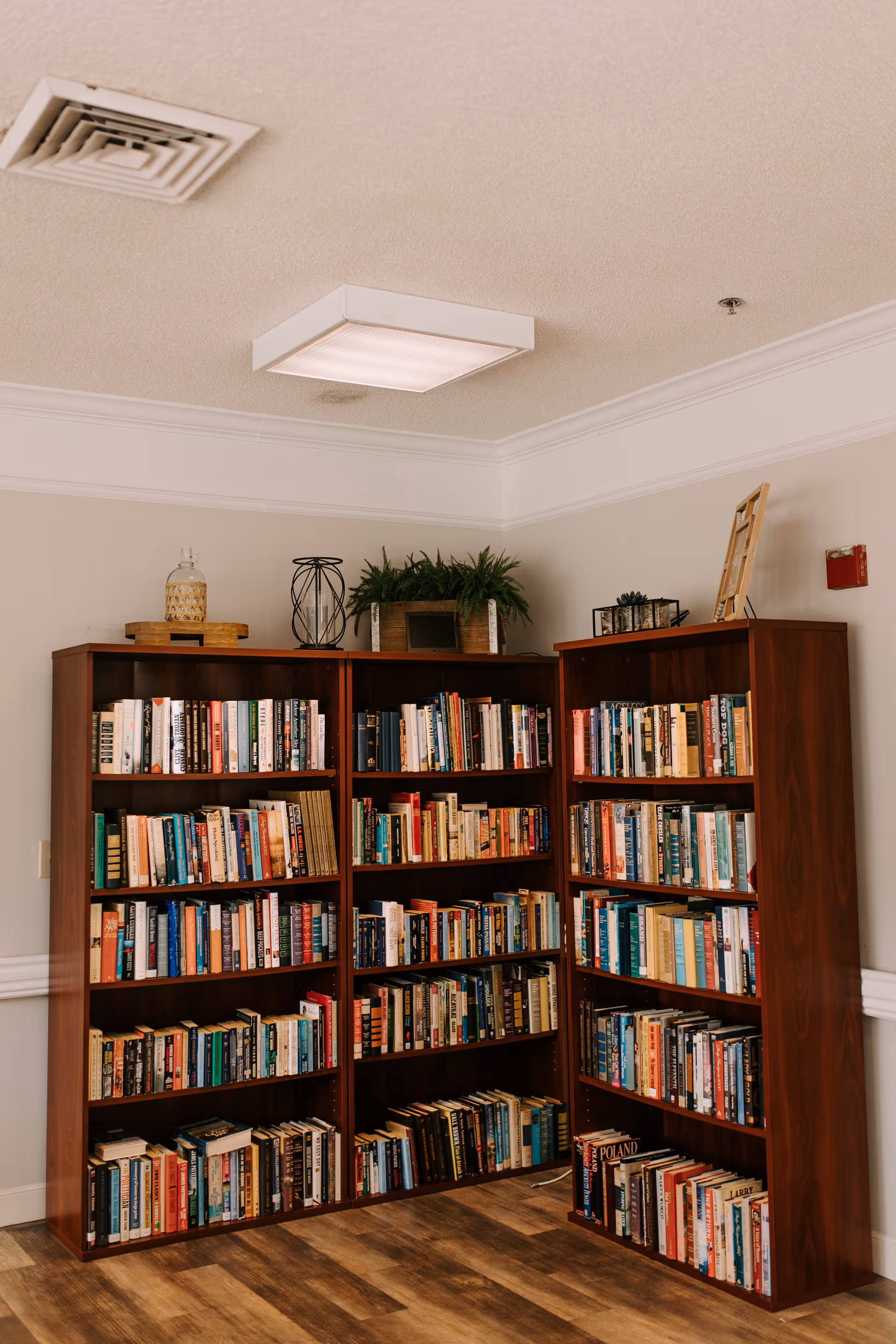Corner of a room with three wooden bookshelves filled with books. On top of the shelves are decorative items including a glass jar, a black metal candle holder, a wooden planter with green plants, and a small framed picture. The room has light-colored walls, a white ceiling with a square light fixture, and wood-patterned flooring.