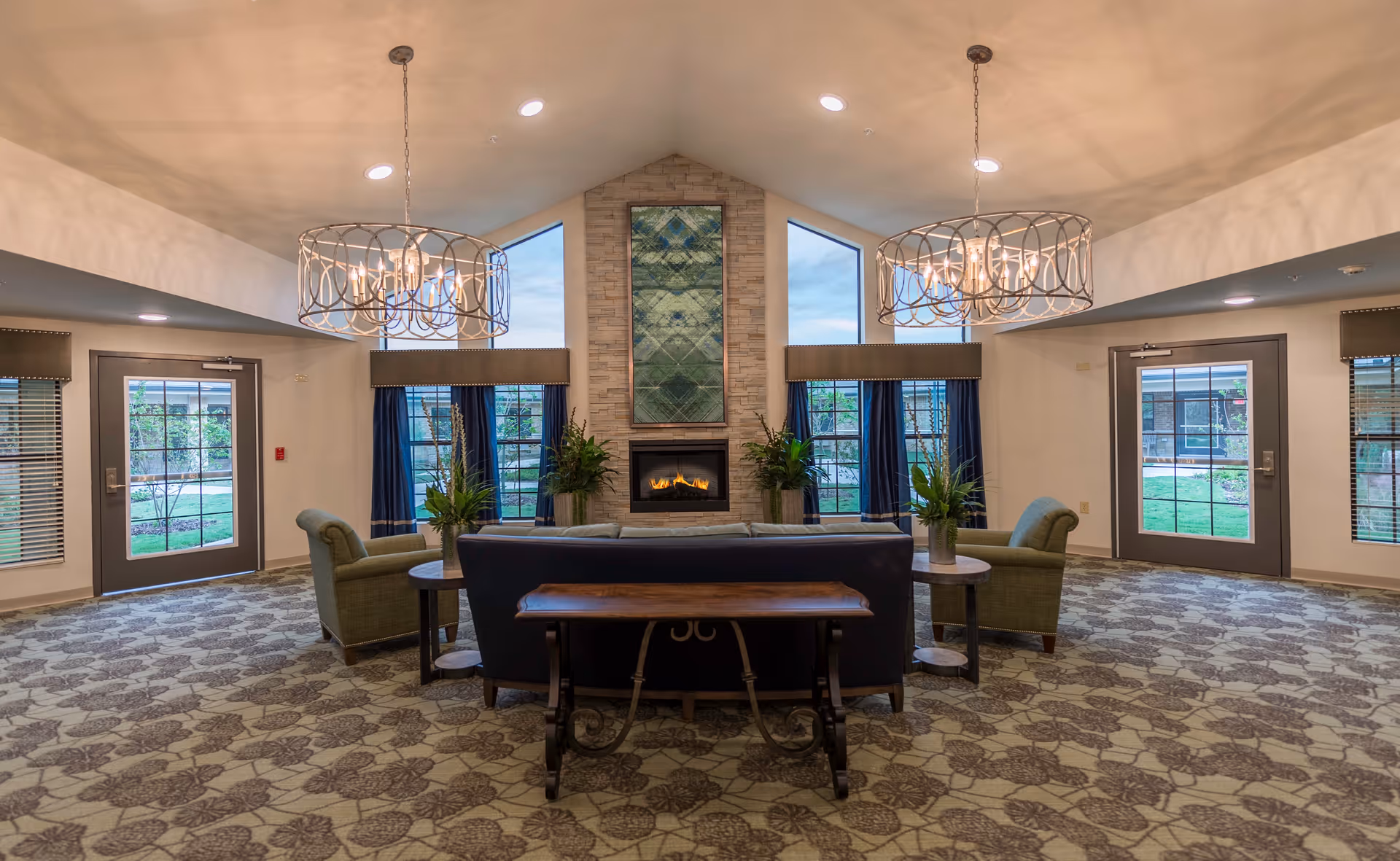 Spacious communal lounge with sofas and armchairs arranged around a central fireplace beneath decorative chandeliers and large windows.