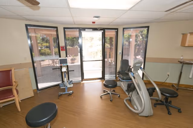A small exercise room with wooden flooring and beige walls featuring large windows and a glass door leading outside. The room contains exercise equipment including a recumbent bike and stools, with a red chair and a small table visible on the sides.