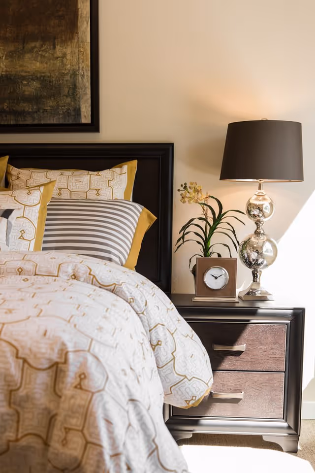 Close-up of a neatly made bed with patterned bedding and pillows in white, gold, and gray tones. Next to the bed is a dark wooden nightstand with two drawers, a silver table lamp with a dark lampshade, a small clock, and a potted plant with green leaves and light-colored flowers. A framed abstract painting hangs on the wall above the bed.