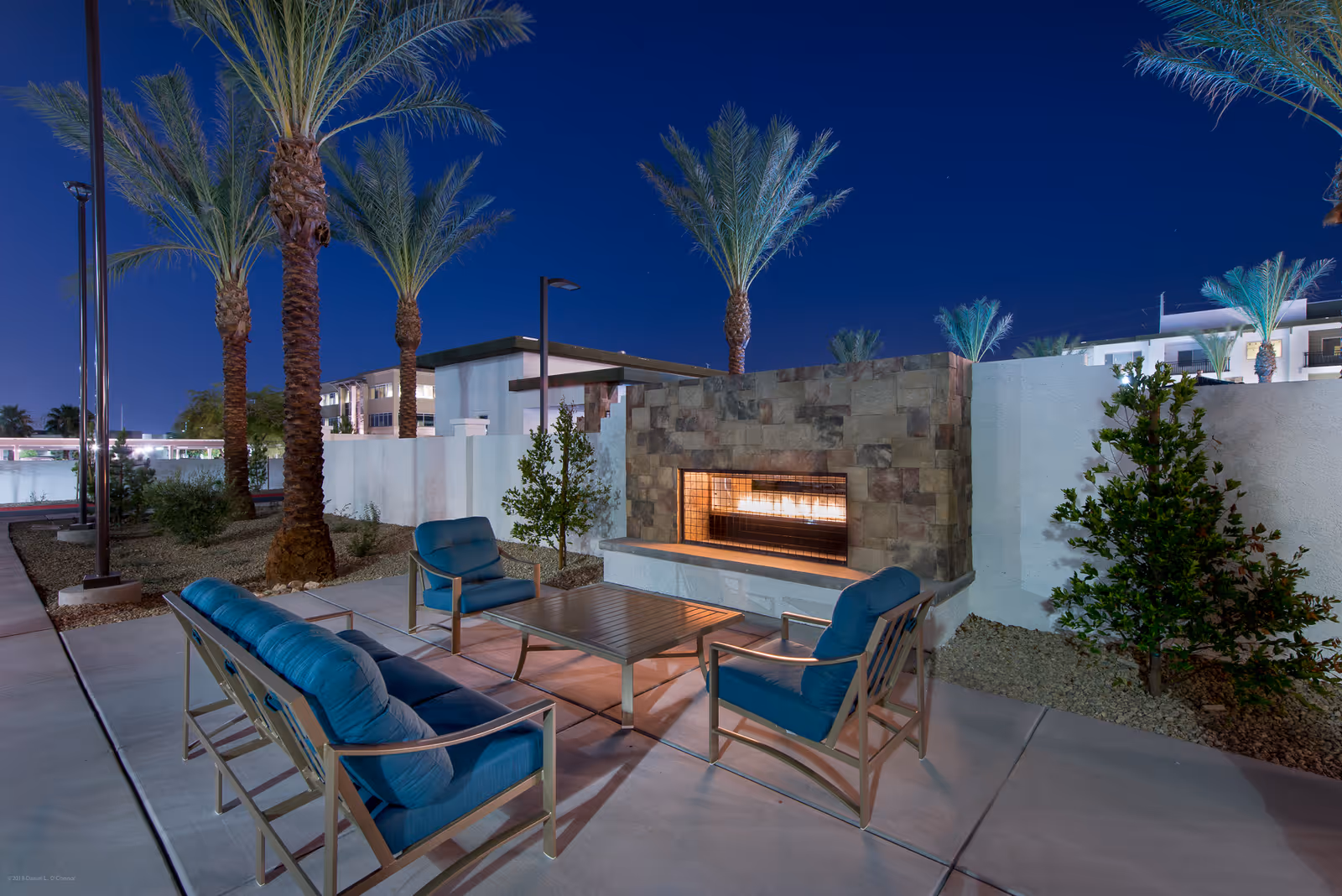 Outdoor seating area at night with blue cushioned chairs and a sofa arranged around a rectangular coffee table in front of a stone fireplace. Tall palm trees and shrubs surround the area, with a clear dark blue sky overhead.