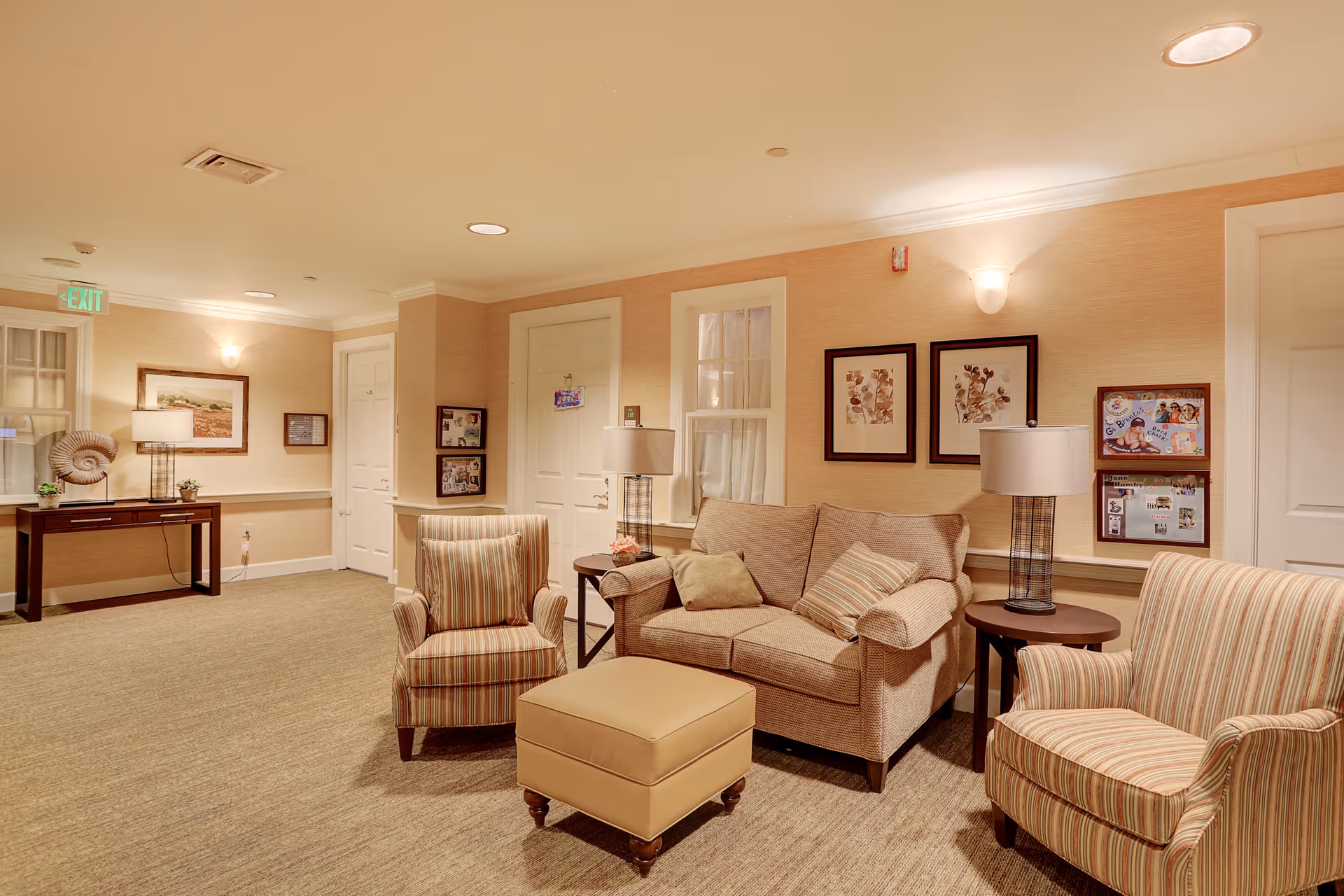 A cozy seating area in a senior living facility with a beige sofa, two striped armchairs, and a matching ottoman. The walls are decorated with framed artwork and photos, and there are two table lamps on either side of the sofa. A console table with a lamp and decorative items is visible in the background near a door with an exit sign above it.