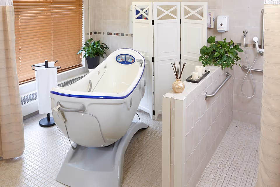 A clean, accessible bathroom featuring a specialized white bathtub with blue trim designed for assisted bathing. The room includes beige tiled walls and floor, a privacy folding screen, green potted plants, a towel stand with a white towel, a soap dispenser, and a handheld showerhead with grab bars for safety.