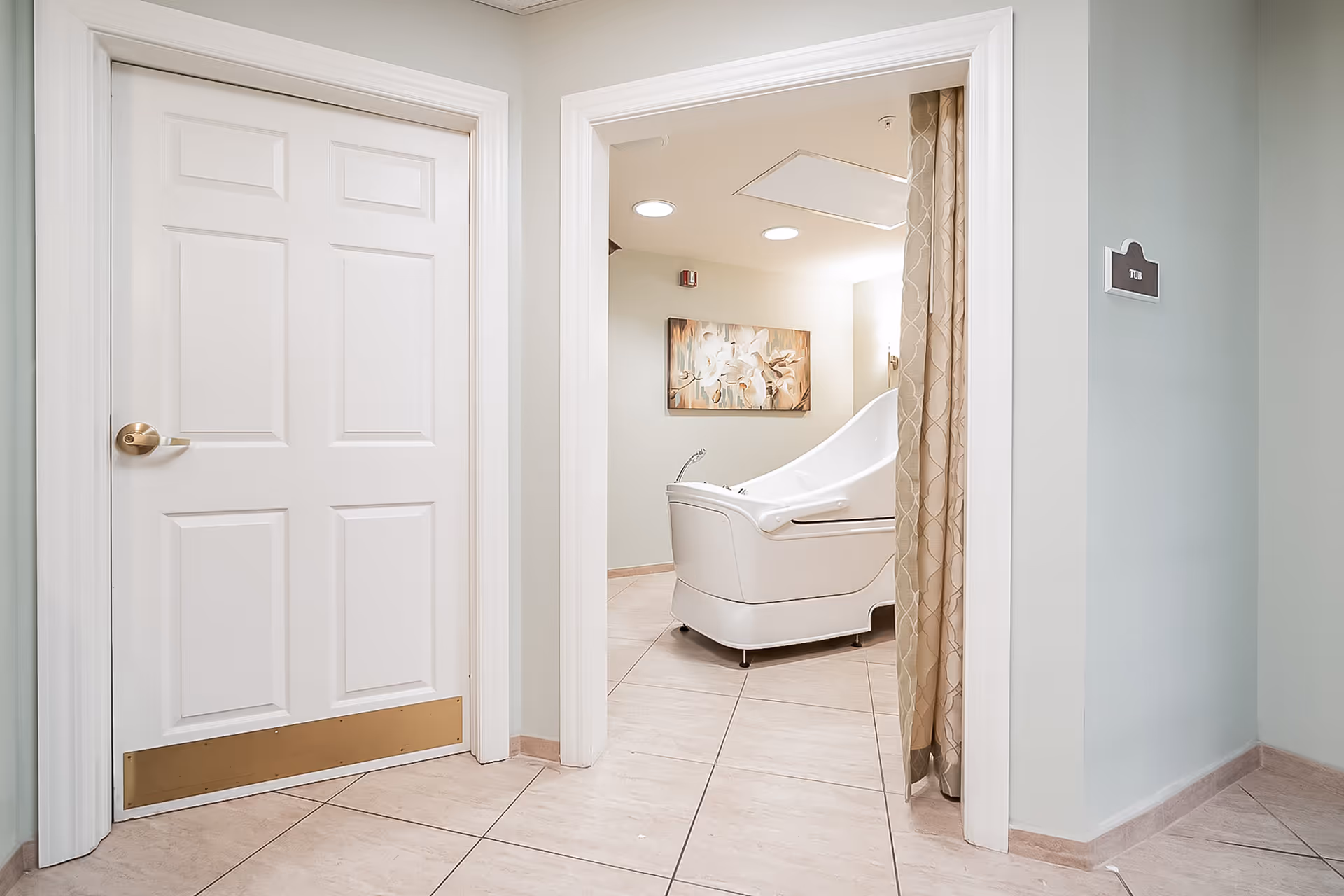 View of a hallway leading to a room with a white therapeutic bathtub. The hallway has beige tiled floors, light-colored walls, and a white door on the left. A curtain partially covers the entrance to the room with the tub. A floral painting hangs on the wall inside the room.