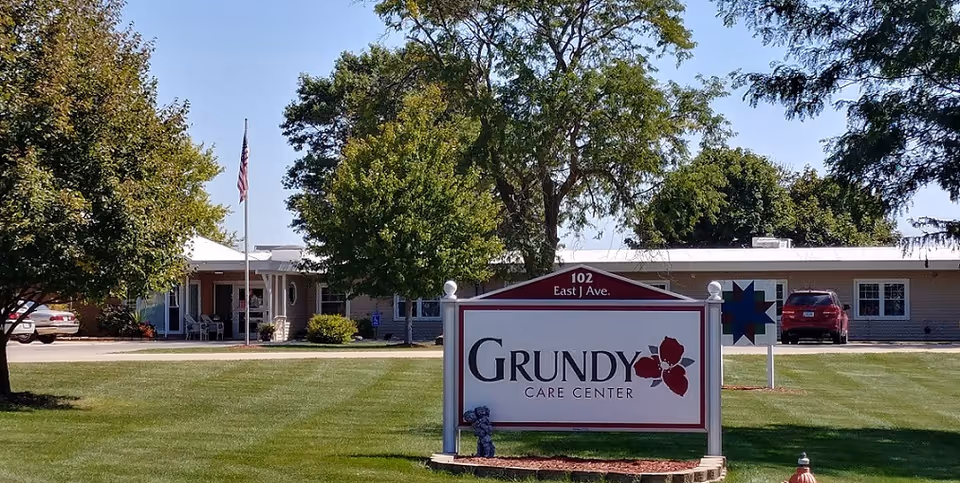 Exterior view of Grundy Care Center building with a large sign in the foreground displaying the facility name and address. The building is a single-story structure surrounded by green grass, trees, and a clear blue sky. There are a few parked cars and an American flag near the entrance.