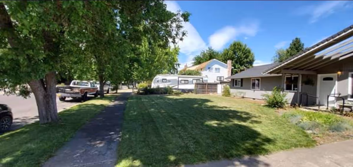 A grassy front yard with a concrete sidewalk leading to a single-story house with a covered porch. There are trees lining the street and a parked truck and trailer visible in the background under a partly cloudy sky.