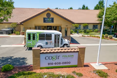 Front entrance of Cogir of Folsom Memory Care with a shuttle parked in front and a large sign displaying the facility name and phone number.
