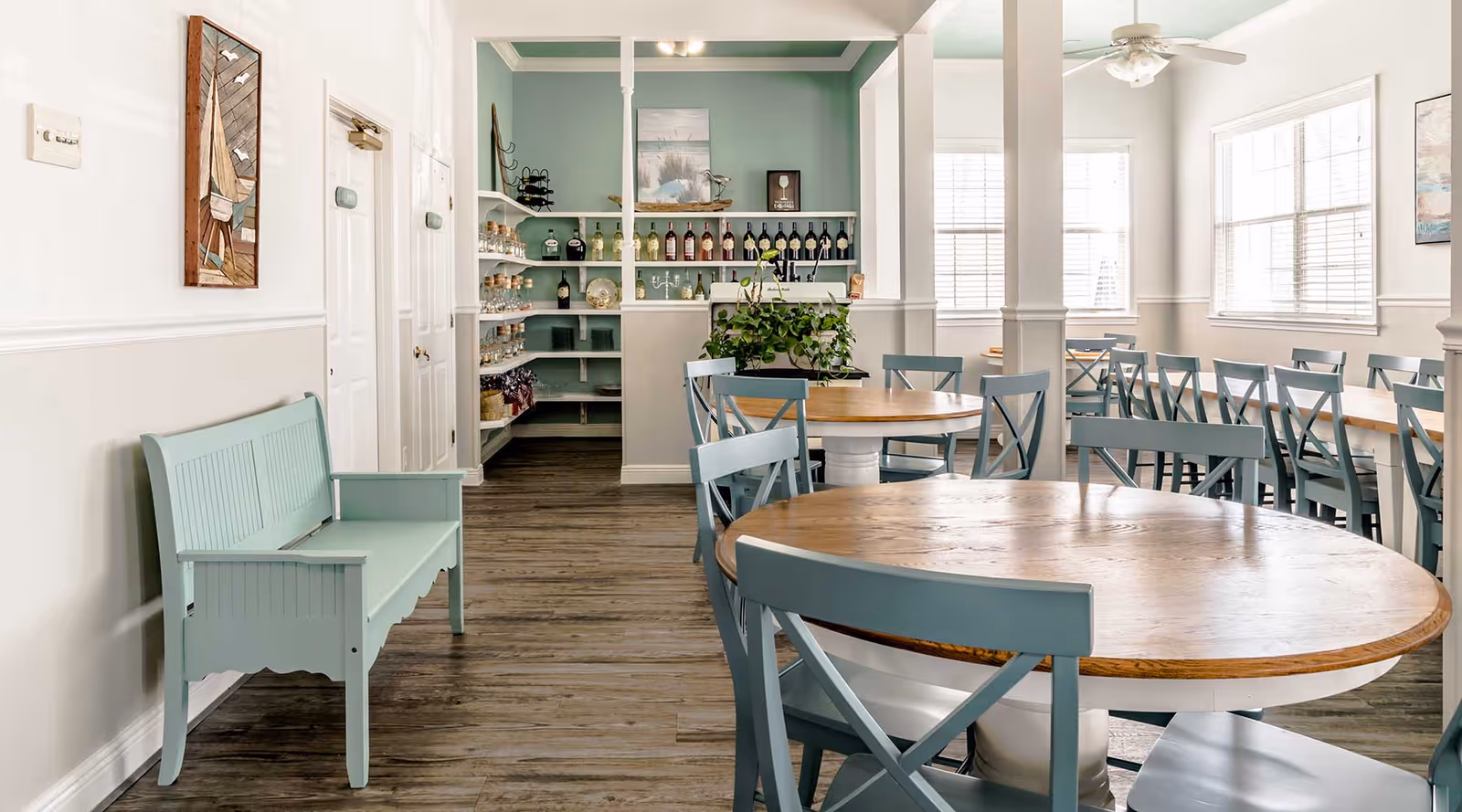 A bright dining room with round wooden tables and light blue chairs. There is a matching light blue bench along the left wall, and a small bar area with shelves holding bottles and glassware in the background. The room has large windows letting in natural light and white walls with light blue accents.