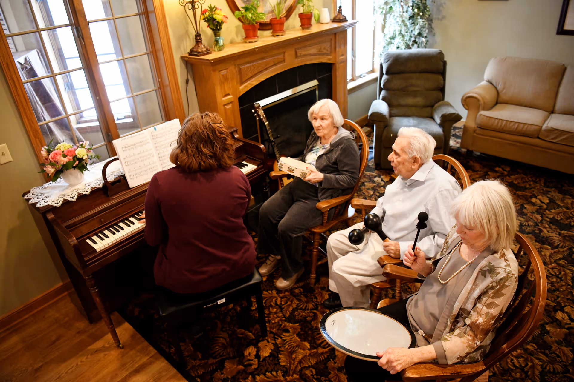 Four elderly residents sit in a cozy living room around a piano while one person plays and others hold percussion instruments.
