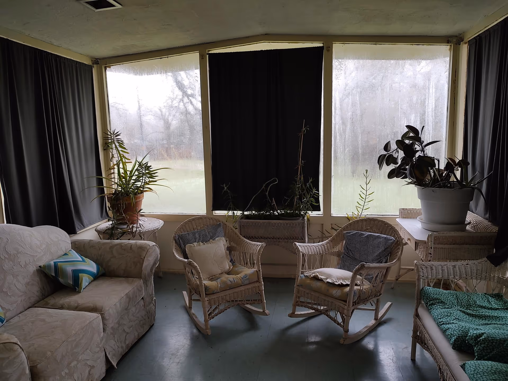 A cozy indoor sitting area with wicker rocking chairs and a loveseat, all with cushions. There are several potted plants on tables and window sills. The windows are large but mostly covered with dark curtains, and the outside view is foggy or rainy.