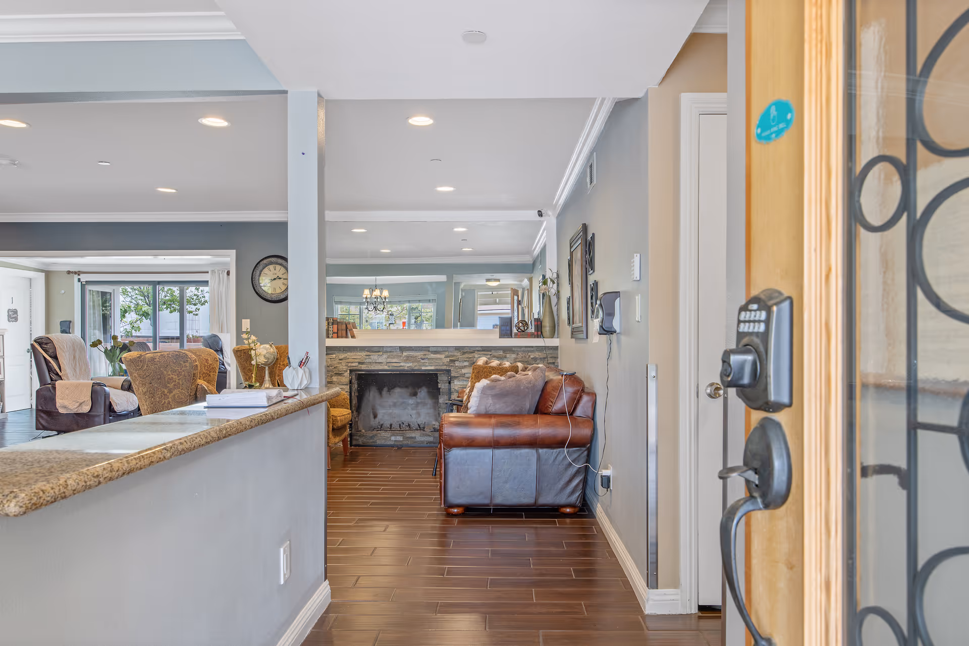 View into a cozy living room area from an open doorway with a keypad lock. The room features a stone fireplace, a brown leather sofa with cushions, a wooden floor, and a granite countertop on the left side. In the background, there are armchairs and large windows letting in natural light.