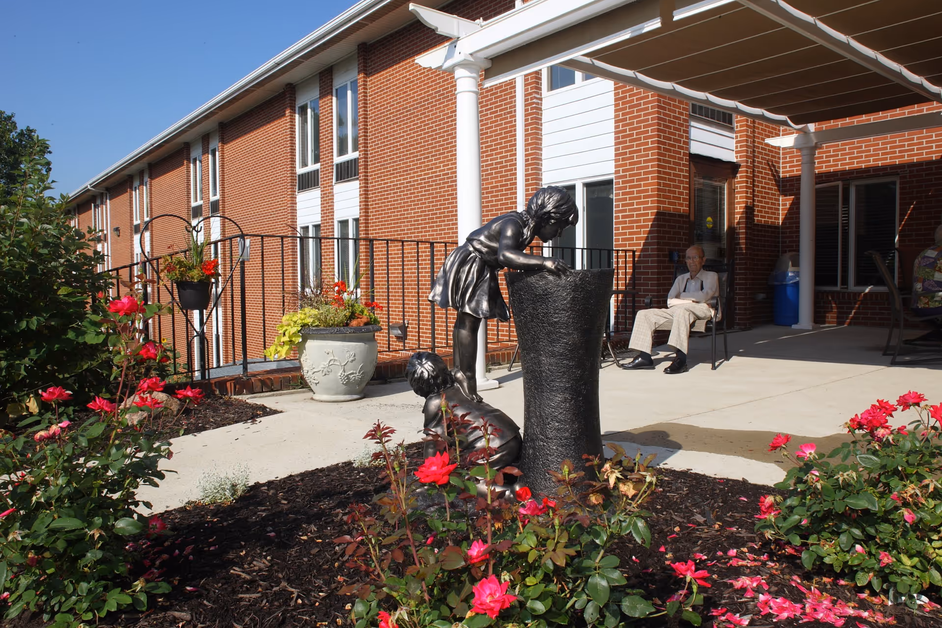 Brick senior living facility entrance patio with flowerbeds, a bronze child fountain sculpture, and a seated resident under a canopy.