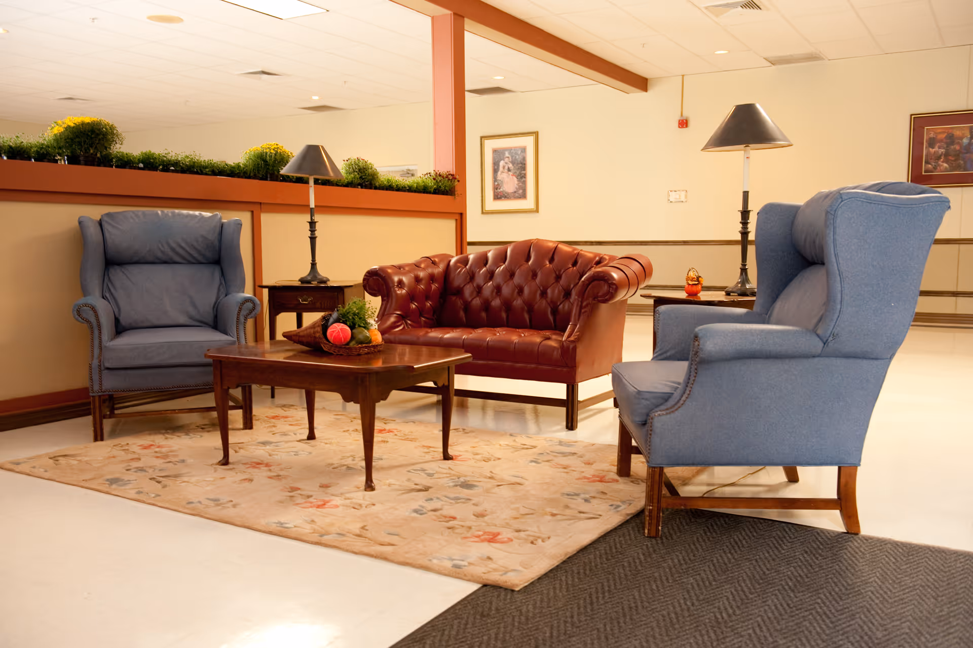 A cozy seating area in a care facility with two blue upholstered armchairs and a brown leather tufted loveseat arranged around a wooden coffee table with a decorative centerpiece. The area is carpeted with a floral rug and has two side tables with lamps. The background shows a beige wall with framed artwork and a planter with green plants on a partition.