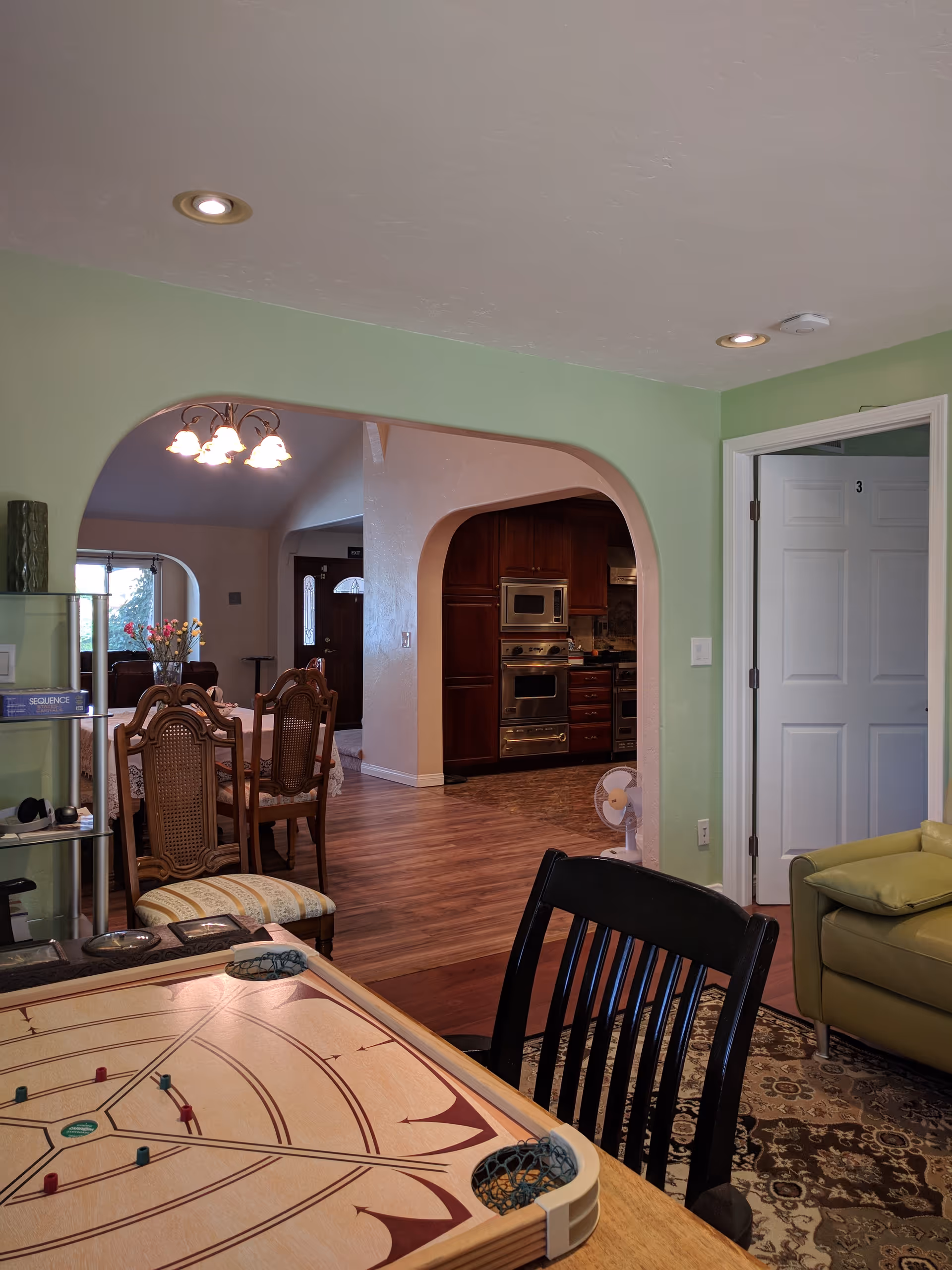 Interior view of a senior living facility showing a game table with shuffleboard in the foreground, a black chair, a green armchair on a patterned rug, and a dining area with wooden chairs and a table with a floral centerpiece. The background features an archway leading to a kitchen with wooden cabinets and stainless steel appliances, and a white door marked with the number 3.