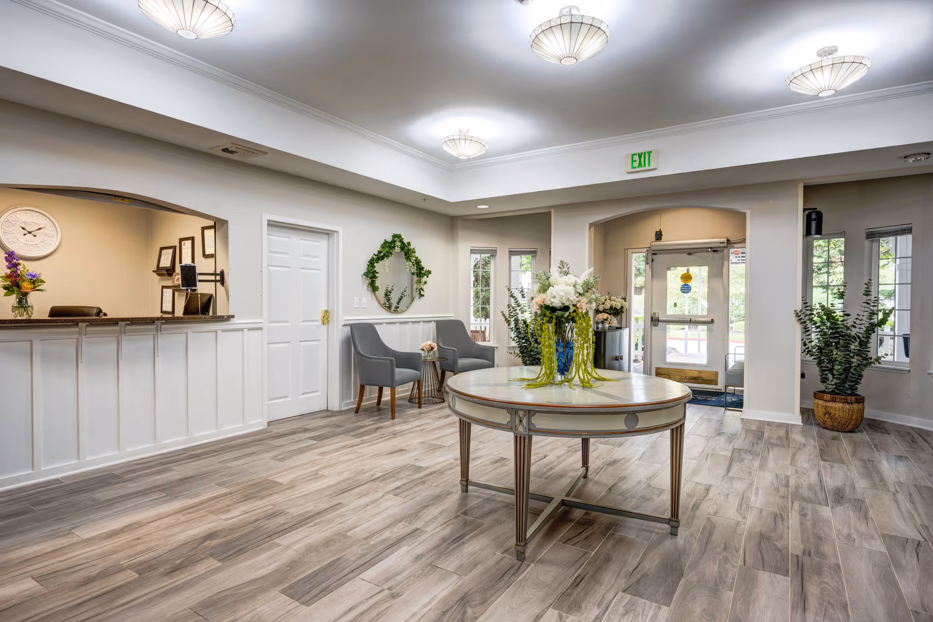 Bright and spacious reception area with a round table in the center decorated with flower arrangements. To the left is a reception desk with a clock and framed certificates on the wall behind it. Two gray chairs with a small side table are positioned against the wall near a decorative wreath. Large windows and glass doors allow natural light to enter the room, and potted plants add greenery to the space. The floor is covered with wood-patterned tiles, and ceiling lights provide additional illumination.