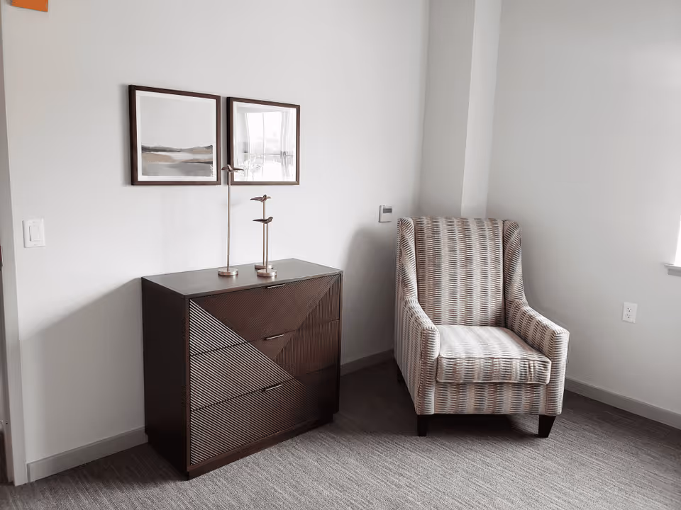 A patterned upholstered armchair beside a dark geometric dresser topped with decorative lamps and two framed prints on a neutral-colored wall.