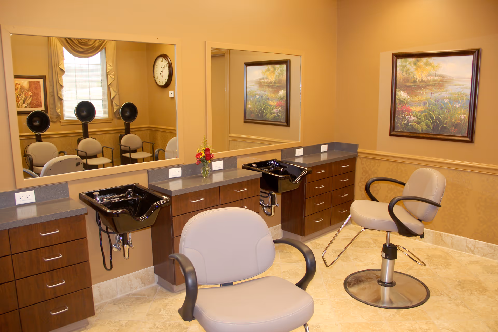 Interior of a hair salon area with two salon chairs, two black hair washing sinks, large mirrors on the wall, wooden cabinets with drawers, a window with curtains, a wall clock, a floral painting, and a small vase with flowers on the counter.