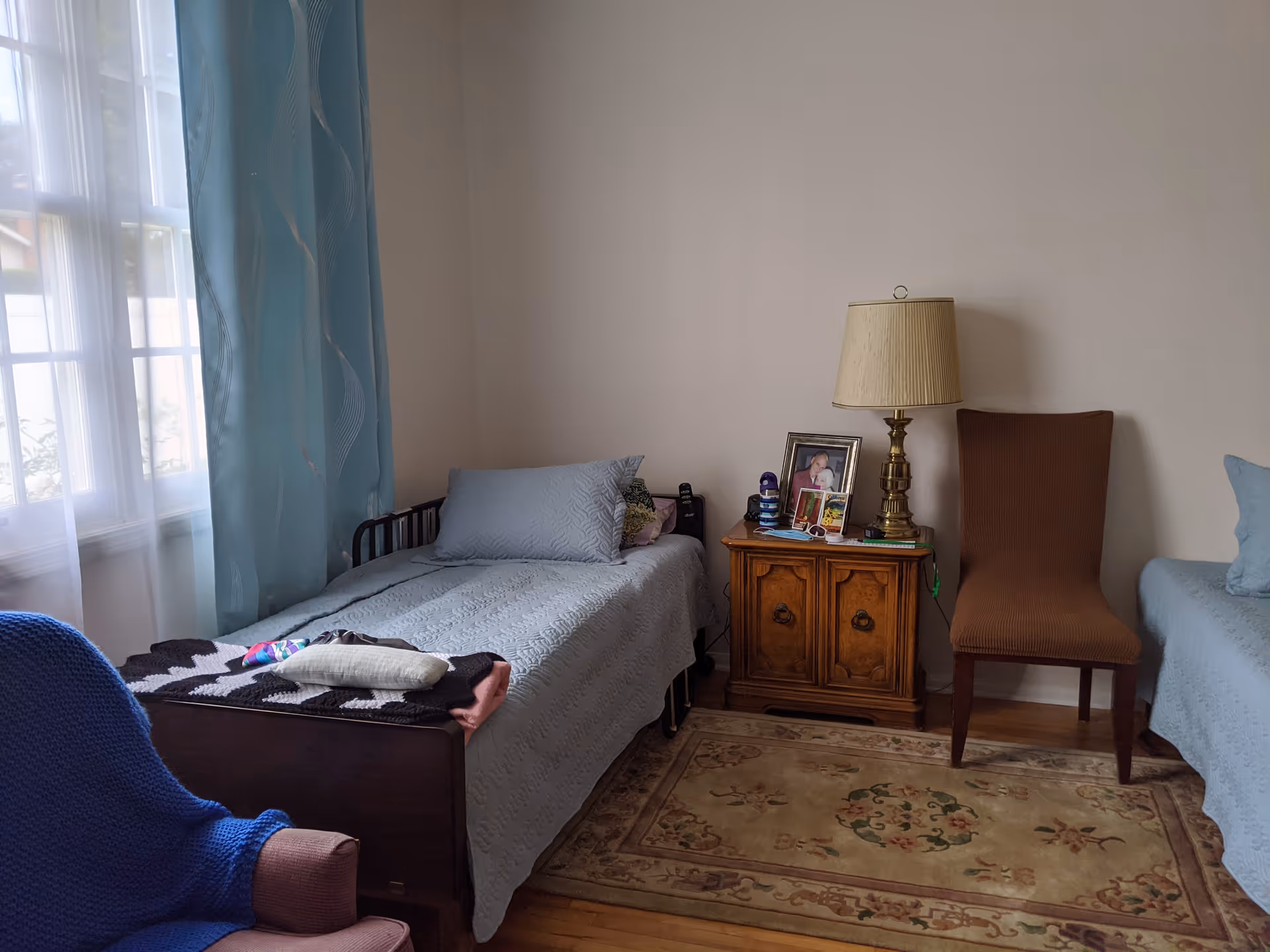 Sunlit bedroom with two twin beds, a wooden nightstand holding a lamp and framed photo, a chair, and a window with blue curtains.