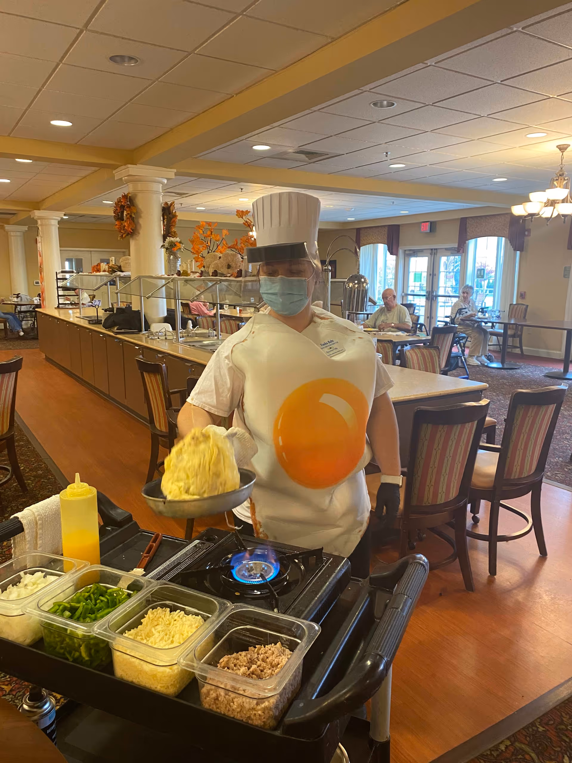 A person wearing a chef's hat, face mask, and a costume resembling a fried egg is cooking an omelette on a portable stove in a dining area. Various ingredients like chopped onions, green peppers, shredded cheese, and cooked meat are in containers on a cart next to the stove. In the background, elderly people are seated at tables in a well-lit dining room with large windows and decorative autumn wreaths on columns.