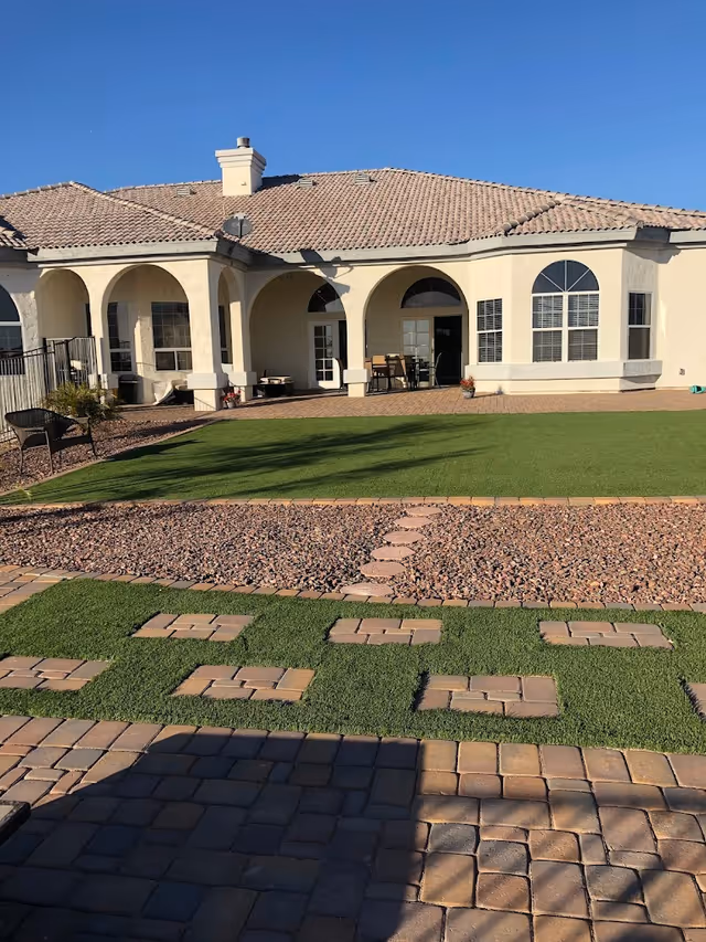 Rear exterior of a stucco assisted living building with arched covered patio, outdoor seating, paved walkways and artificial lawn under a clear blue sky.