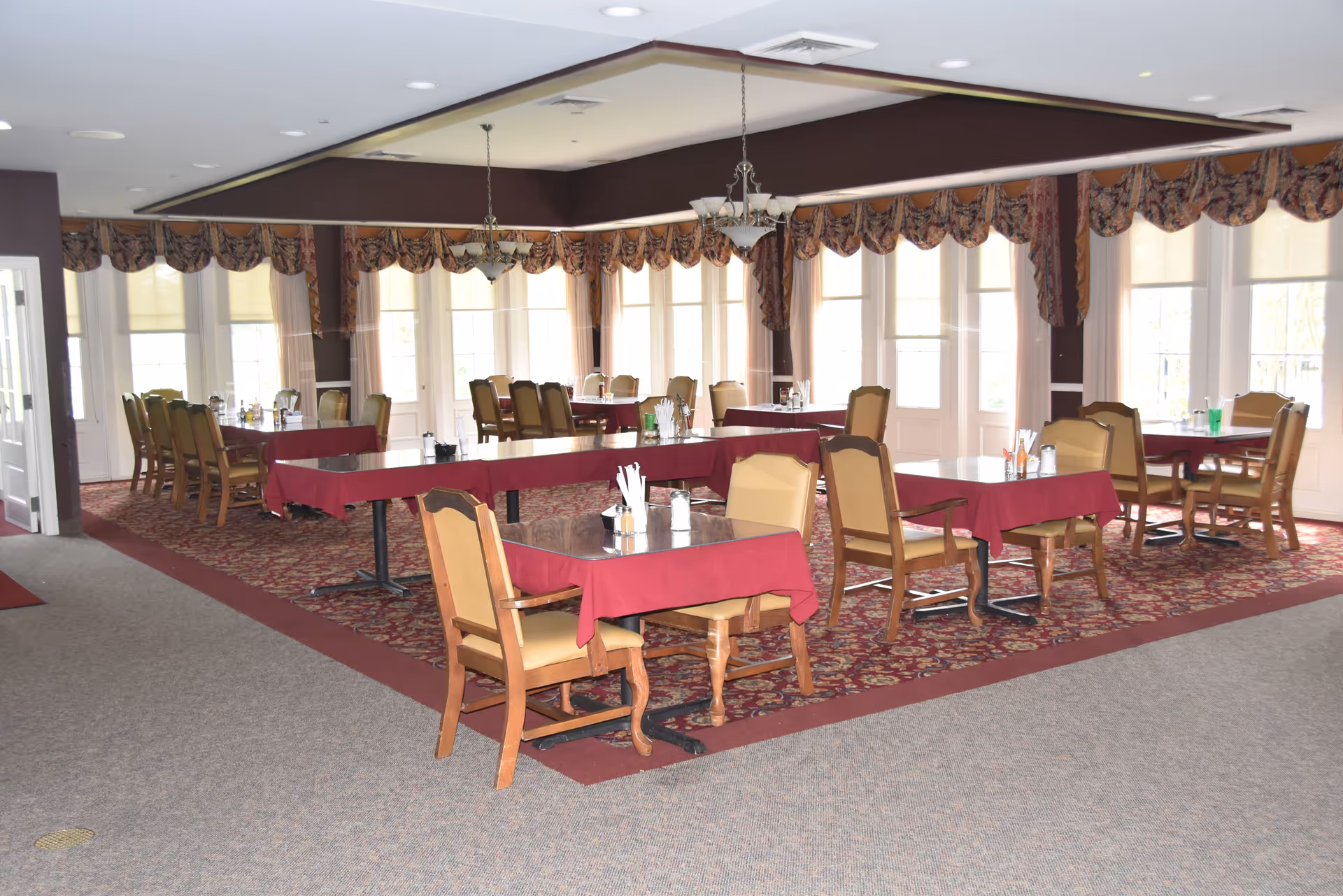 Spacious assisted living dining room with multiple tables covered in red tablecloths and wooden chairs near large windows.