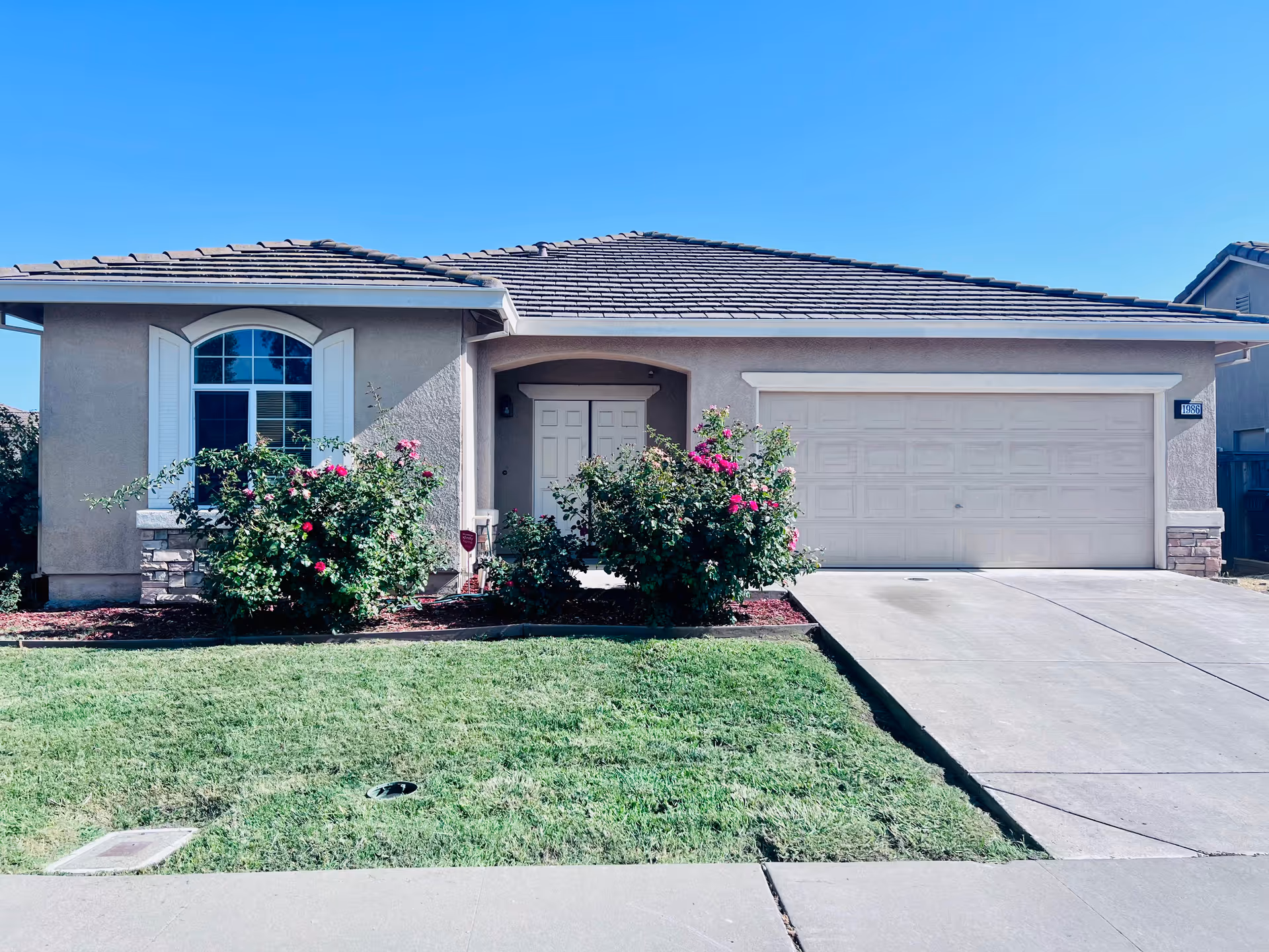 Front exterior view of a single-story house with a tiled roof, a double garage door, a front door with an arched entryway, a large window with white shutters, and a well-maintained lawn with flowering bushes in front.