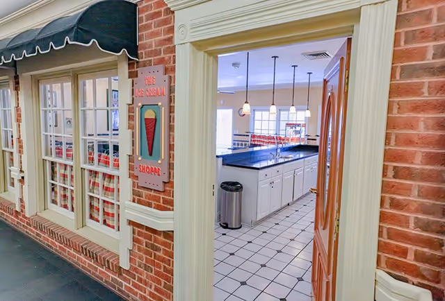 View through an open door into a bright kitchen area with white cabinets, a black countertop, hanging pendant lights, and a trash can. Outside the door is a brick wall with windows covered by red and white checkered curtains and a sign that reads 'The Ice Cream Shoppe' with an ice cream cone illustration.