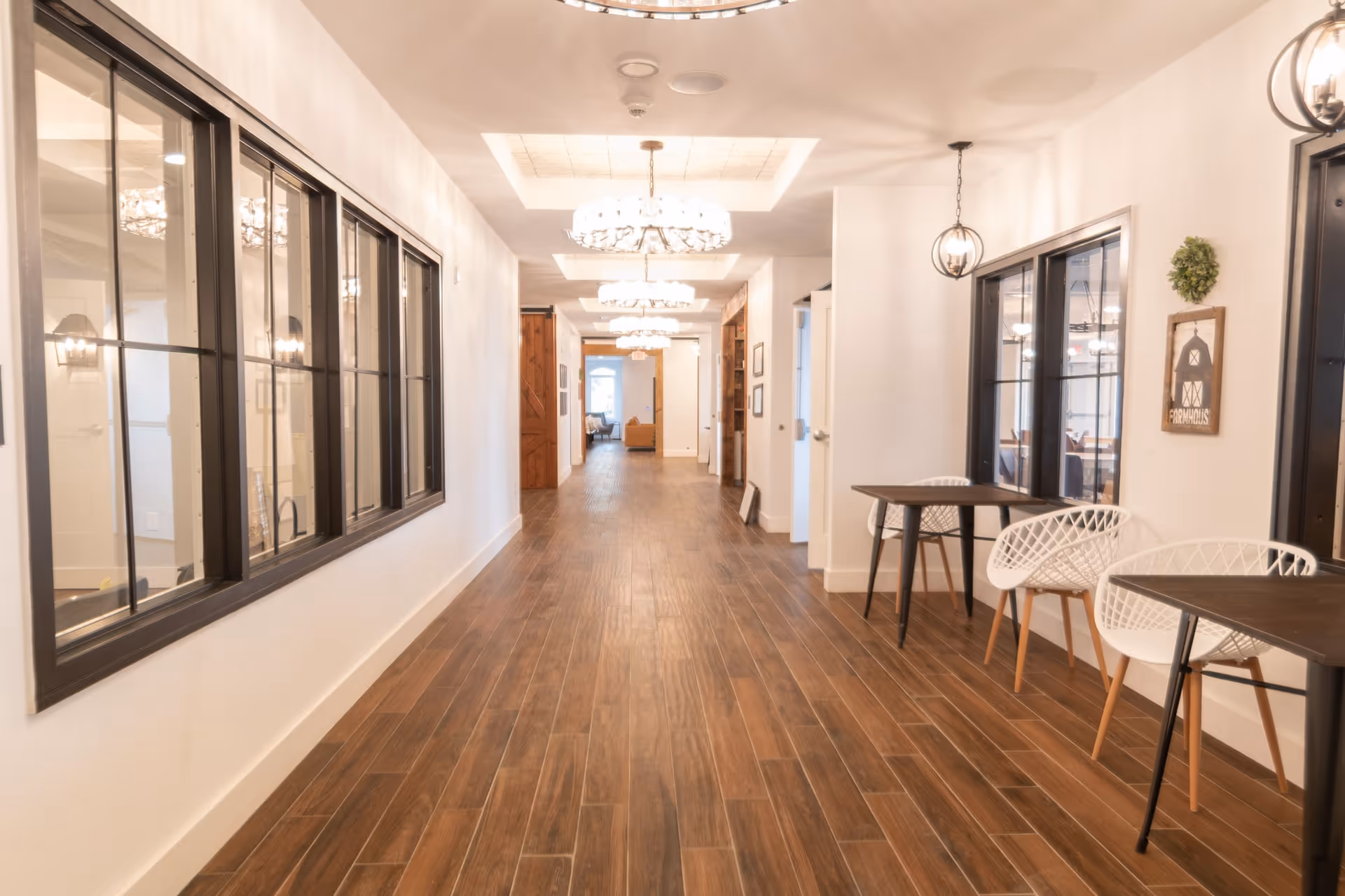 A bright, spacious hallway with wooden flooring and white walls. The hallway features large windows on the left side and small tables with white chairs on the right side. Elegant chandeliers hang from the ceiling, illuminating the corridor. At the far end, there is a seating area with a couch visible.