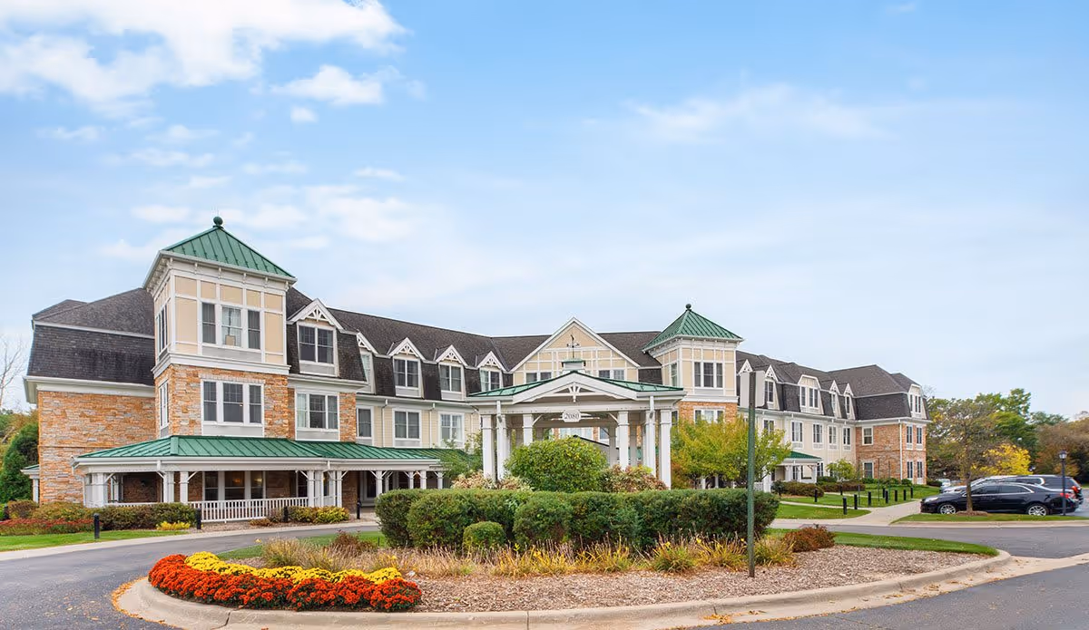 Exterior view of a large senior living facility building with beige and stone walls, green roofs, and multiple windows. The entrance features a covered driveway with white pillars. There is a landscaped roundabout with bushes and colorful flowers in front of the building, and several parked cars are visible on the right side under a partly cloudy sky.