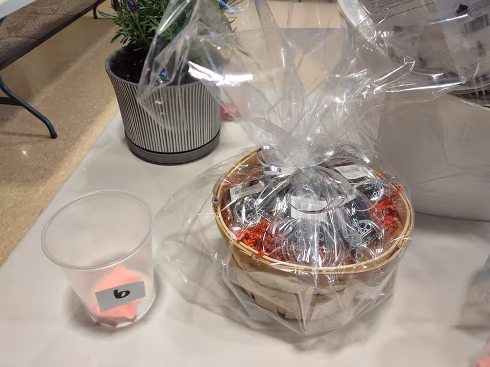 A small gift basket wrapped in clear plastic containing several small items, placed on a table next to a small potted plant and a clear plastic cup with a number 6 label inside.
