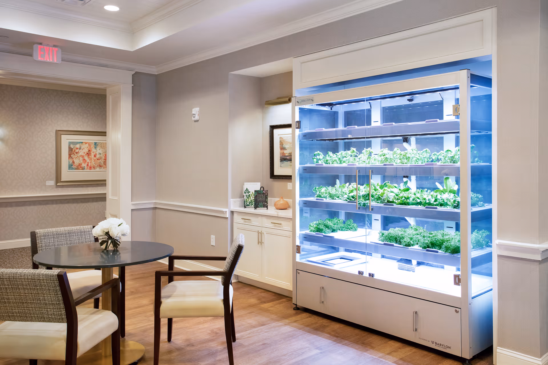 A bright indoor space featuring a round table with four chairs around it, a small vase with white flowers on the table, and a large illuminated indoor garden unit with multiple shelves of green plants. The room has light-colored walls, wood flooring, and framed artwork on the walls.