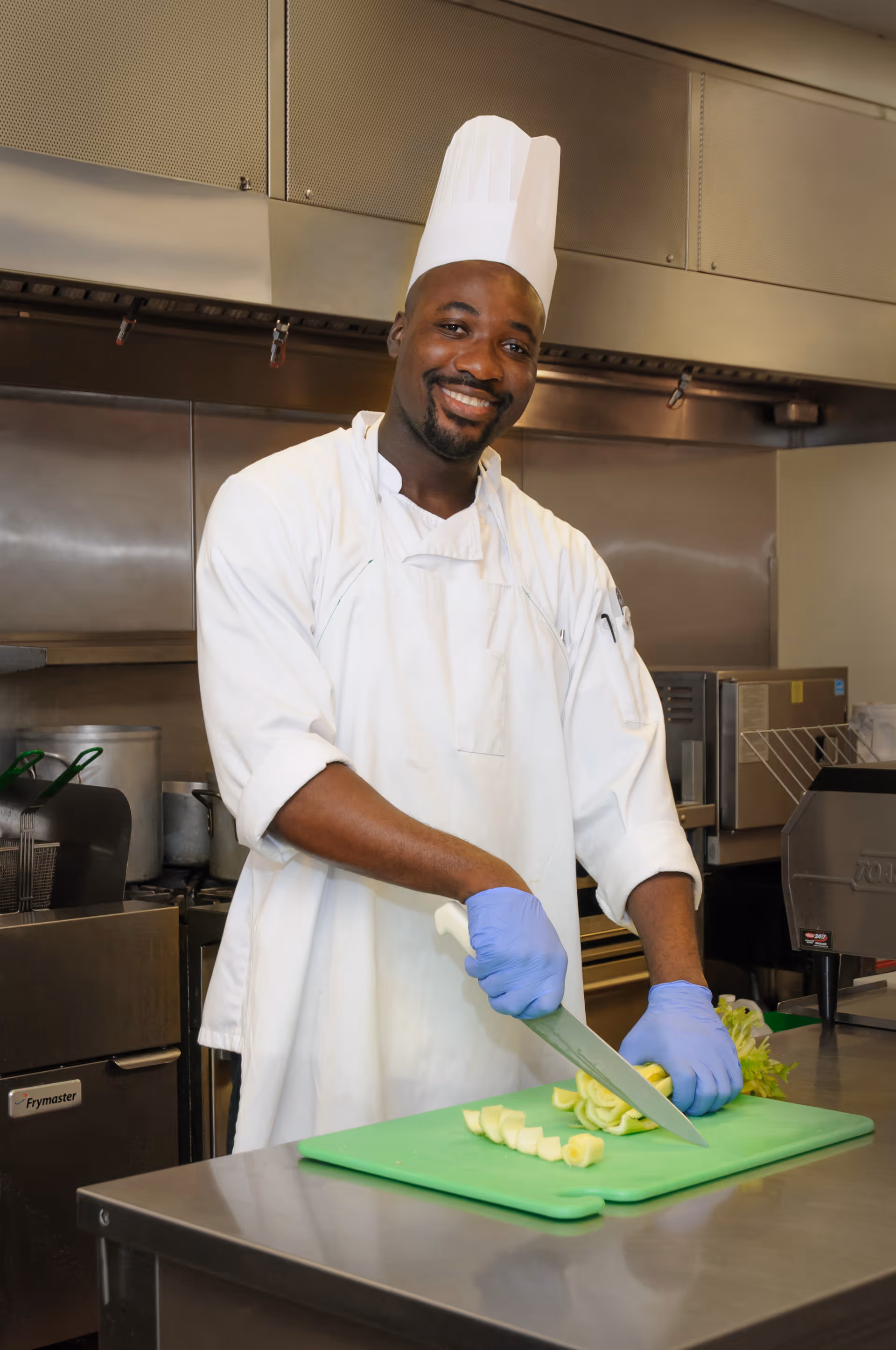 A smiling chef wearing a white uniform, chef hat, and blue gloves is slicing vegetables on a green cutting board in a commercial kitchen.